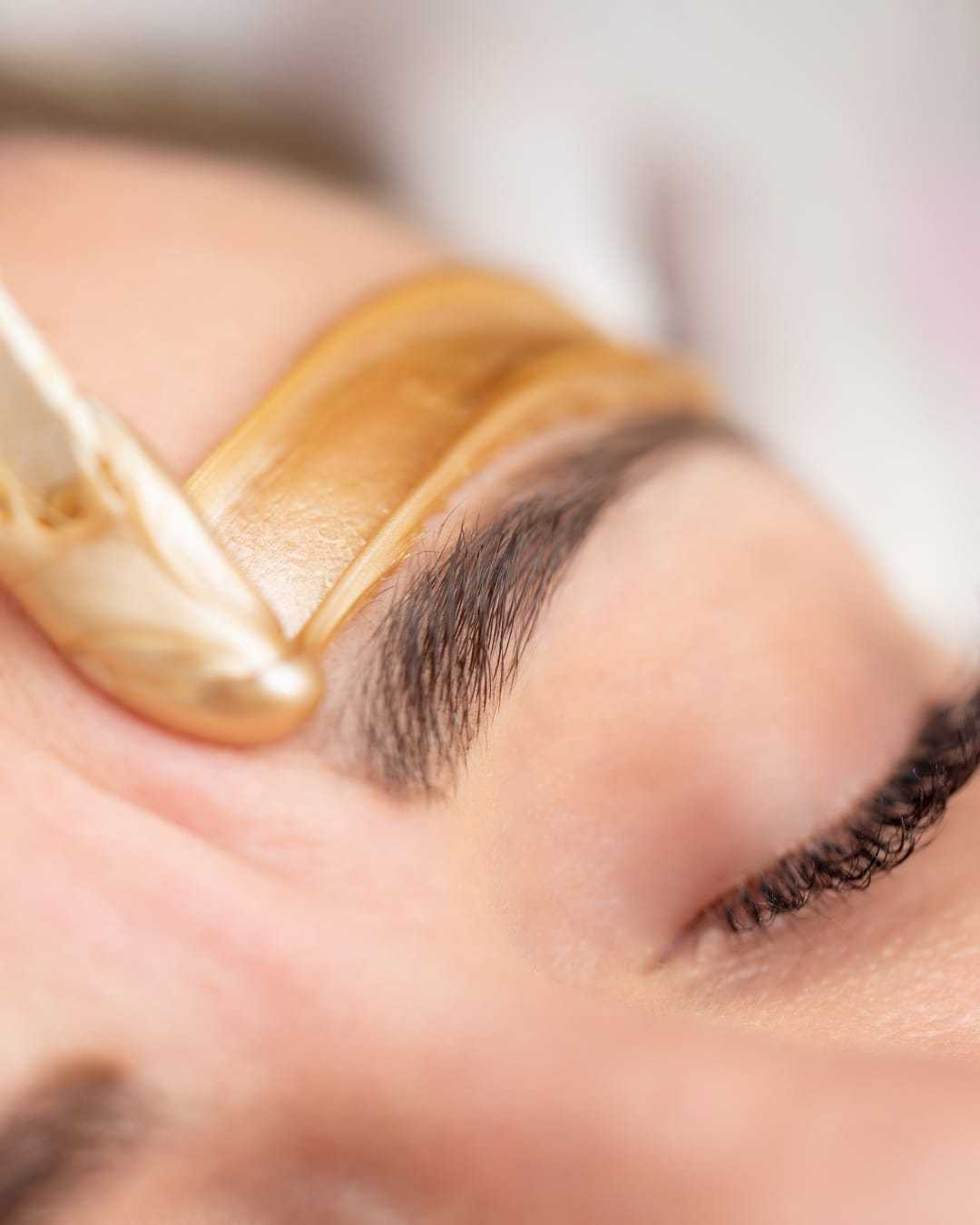 Eyebrow being shaped with a gold wax strip, close-up view.